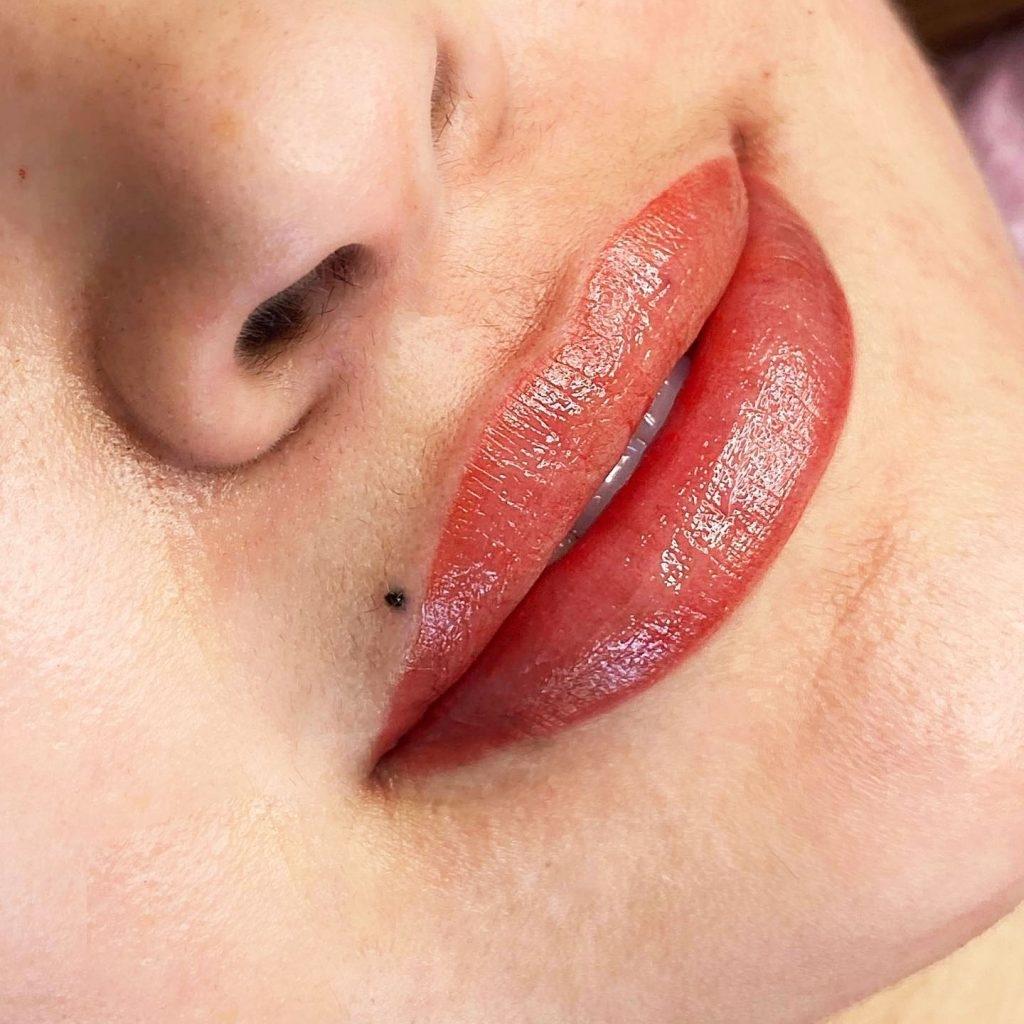Close-up of a person's lips with an even coral-red tint and glossy finish, a small dark mole beside the upper lip.