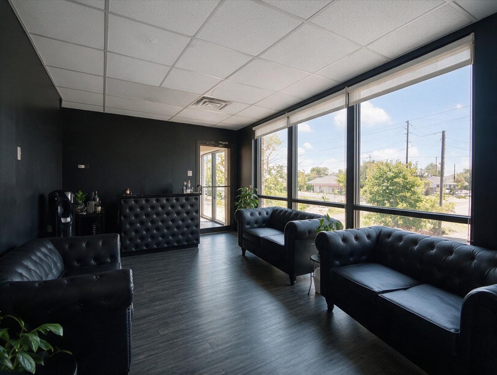 Empty waiting room with black walls, tufted black leather sofas, a padded counter and large windows letting in daylight.